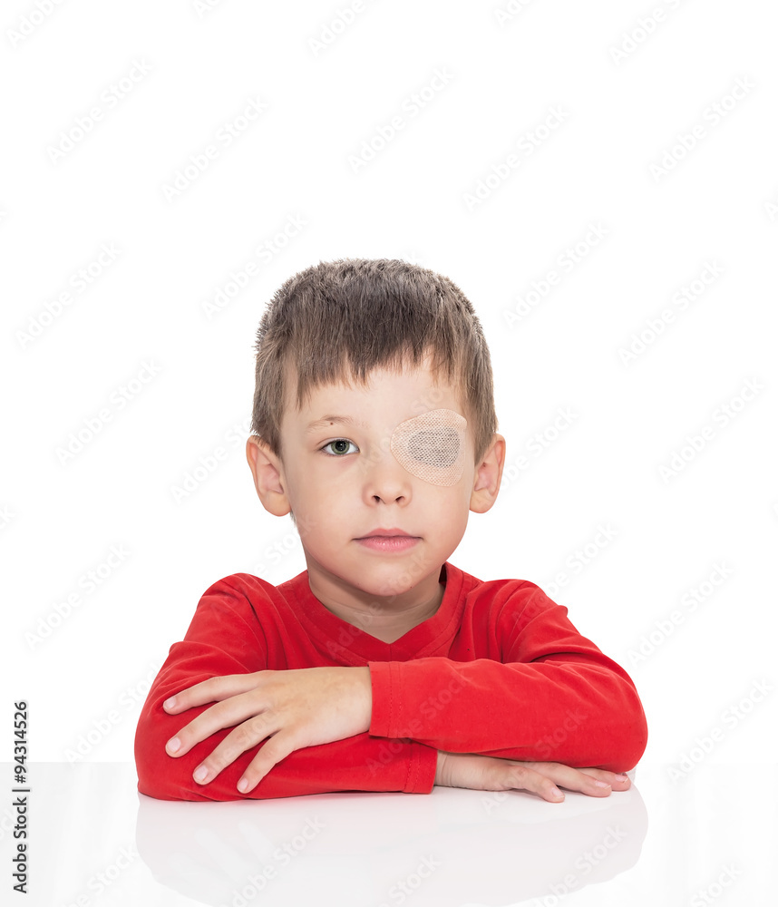 The five-year-old boy sits at a white table, the left eye is stuck with a plaster, sight (vision) stimulation, treatment, load of the right eye, isolated on a white background The five-year-old boy sits at a white table, the left eye is stuck with a plaster, sight (vision) stimulation, treatment, load of the right eye, isolated on a white background