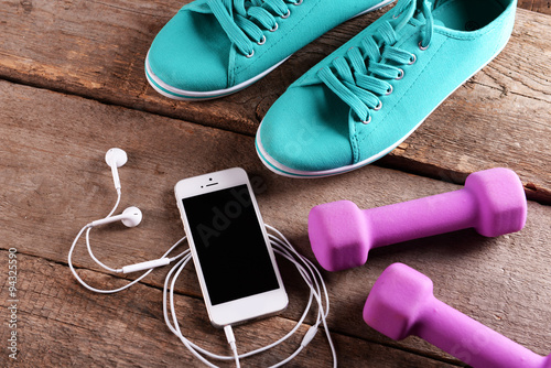 White cellphone with headphones and pink dumb bells and gumshoes on wooden background