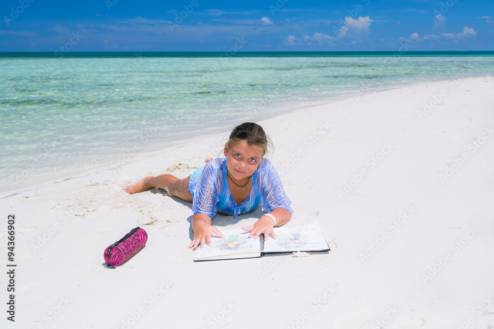 happy charming pretty little girl, lying down on a sunny gorgeous beach and practicing  how to draw in her sketch book