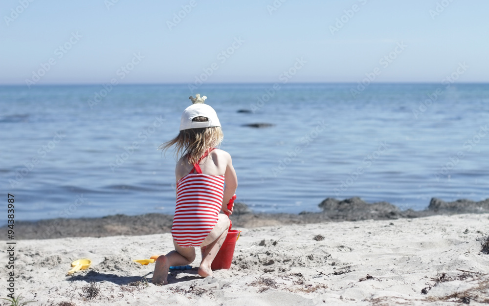 a girl in striped swimming suit sits with her back to the viewer and plays in white sand beach, sea in on a horizon.