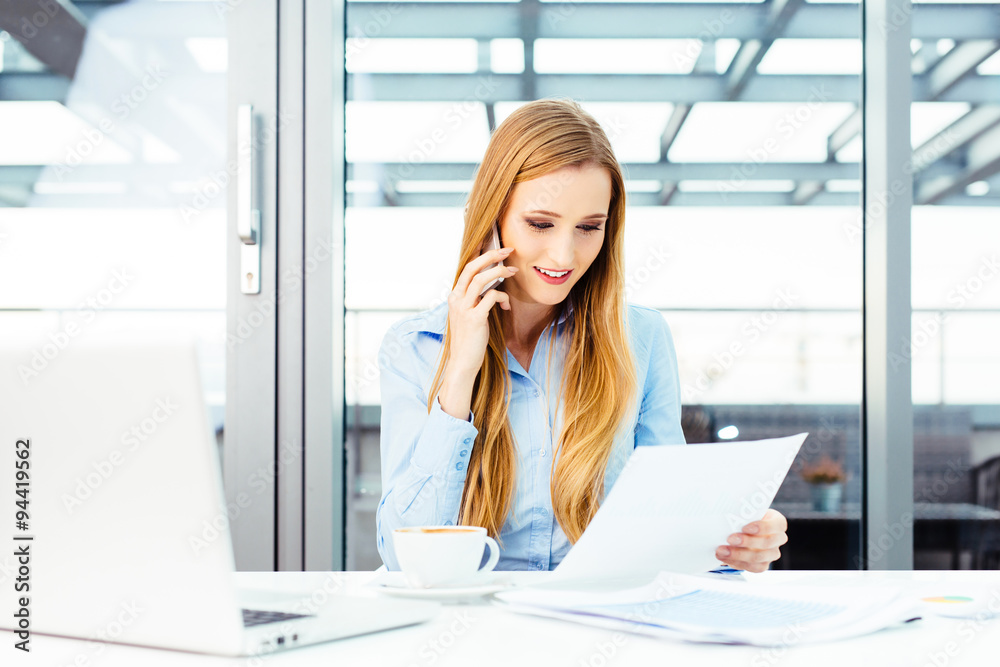 Blonde business woman talking on a mobile sitting at desk with open laptop Blonde business woman talking on a mobile sitting at desk with open laptop