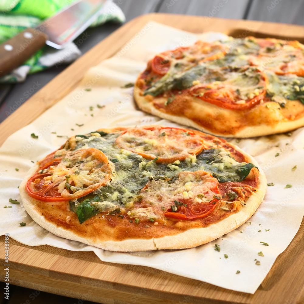 Homemade spinach and tomato pizza on baking paper on wooden board, photographed with natural light (Selective Focus, Focus one third into the first pizza)