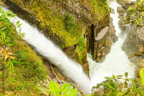 The Pailon del Diablo waterfall plunges through a narrow moss-covered rock canyon creating white frothy water framed by green foliage near Banos, Ecuador, a wild natural wonder.