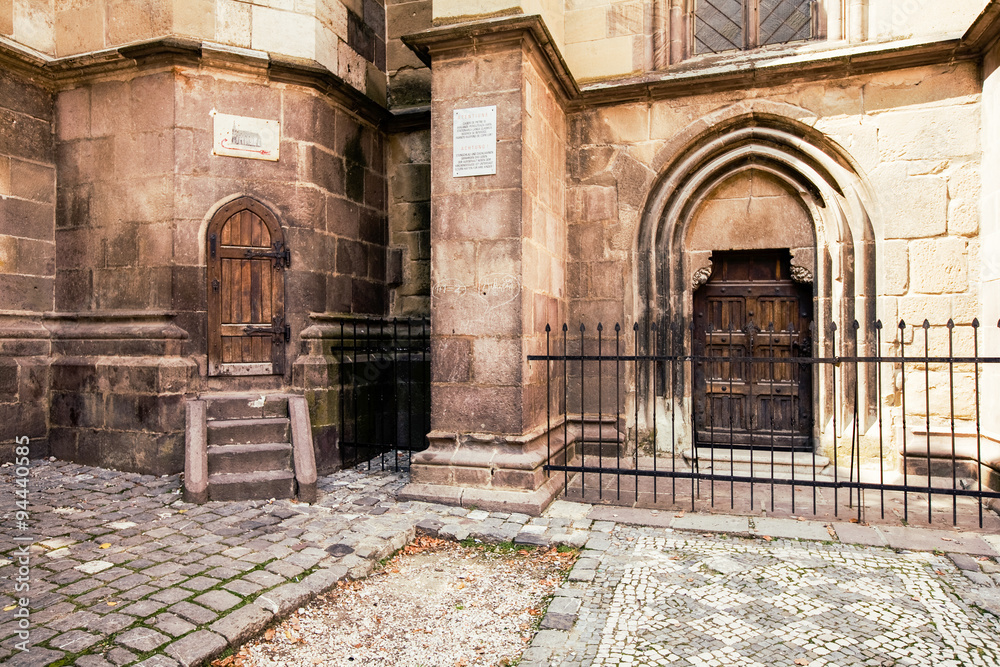 Fototapeta premium A close-up of the ancient Gothic stone facade of the Black Church in Brasov, Romania, a weathered wooden door in a pointed arch behind an iron fence and cobblestone courtyard.
