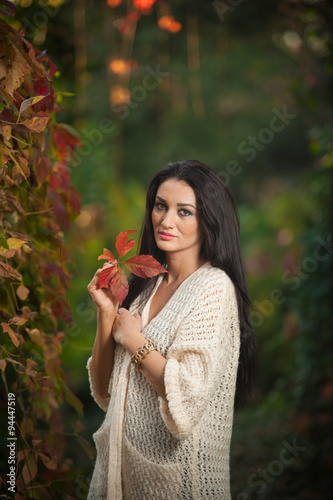 Beautiful woman in white posing in autumnal park. Young brunette woman spending time in autumn near a tree in forest. Long dark hair attractive woman smiling with faded leaves around her
