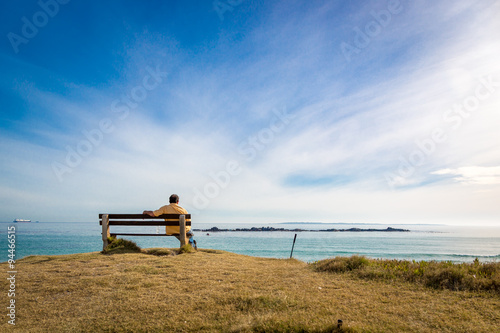 Lonely man sitting a public seat in front of a nice beach in a sunny blue sky day. Concept of dreaming, thinking, planning, looking forward...