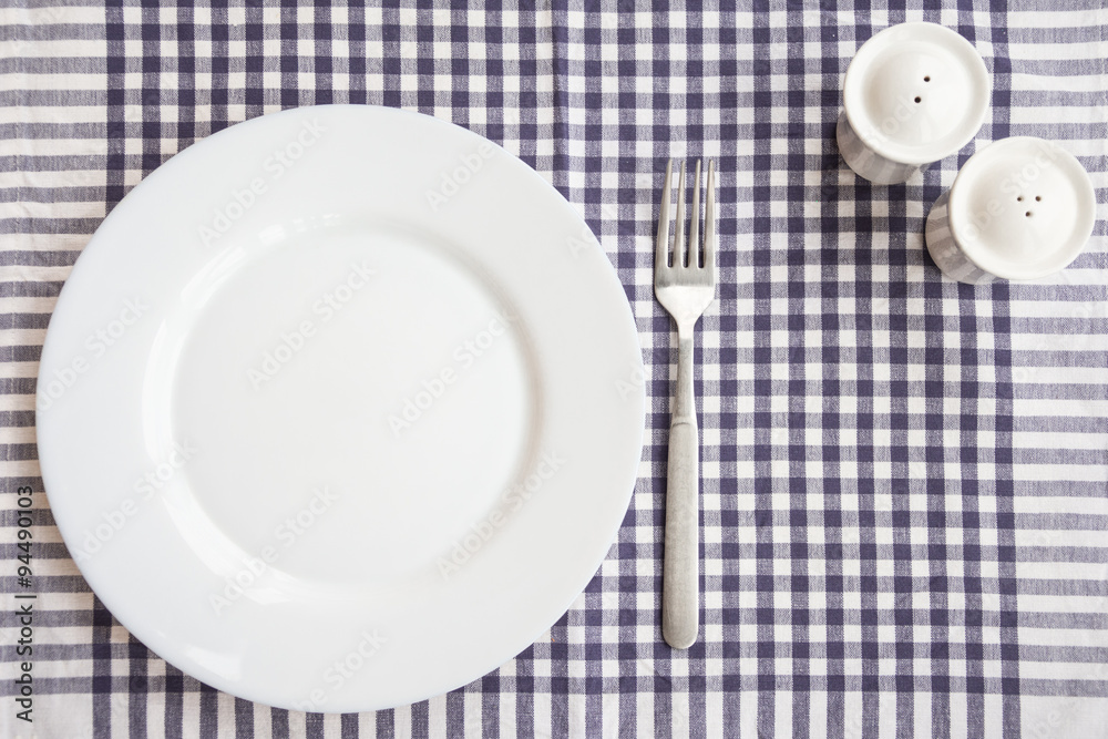 Empty white plate with a fork, pepperbox, salt-cellar, on a checkered tablecloth Empty white plate with a fork, pepperbox, salt-cellar, on a checkered tablecloth