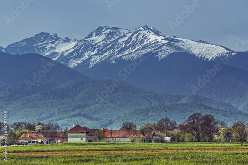 Peaceful rural scenery with traditional Romanian village at the base of snowy Fagaras mountains during spring season.