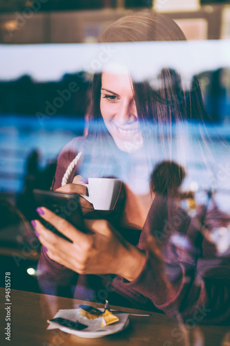 Young woman in cafe  drinking coffee and using her mobile phone.   Seen through the window