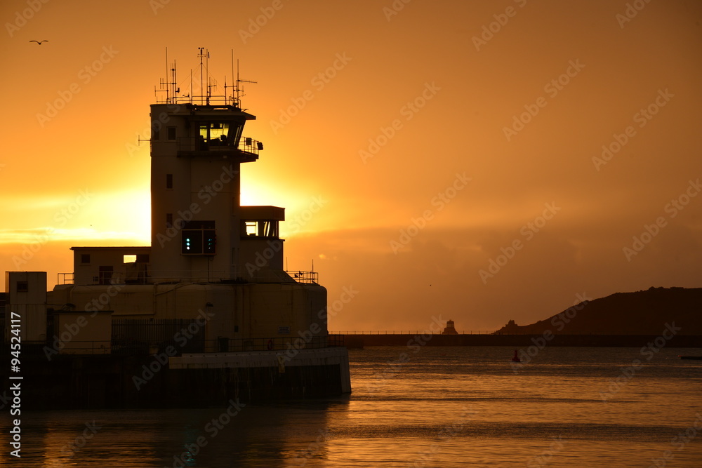 St.Helier harbour, Jersey, U.K. Telephoto image of the sun setting behind a port traffic control tower at high tide in Autumn.