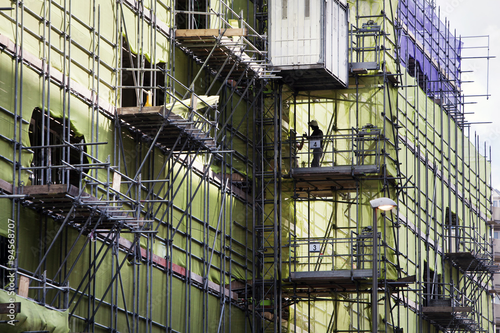 Scaffolding with green and purple protection nets. Two workers discussing in the background.