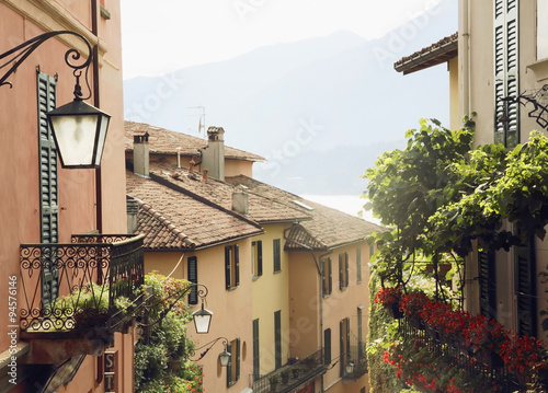 mediterranean buildings with tile roofs, balconies, shutteres, grapes leaves and flowers. Mountains and lake Como on background.  photo