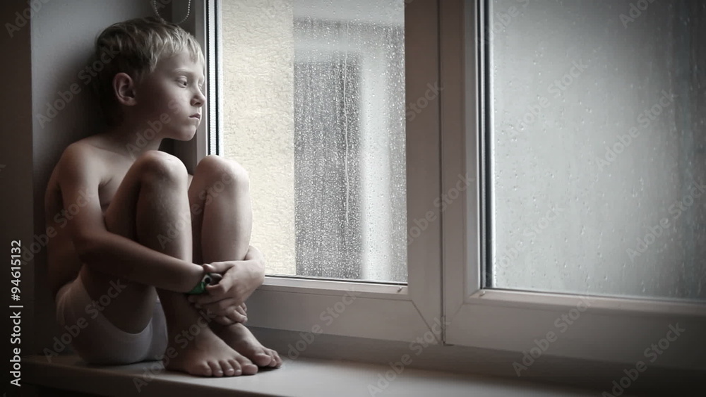 Moody little boy sitting on the windowsill watching raindrops go down the window