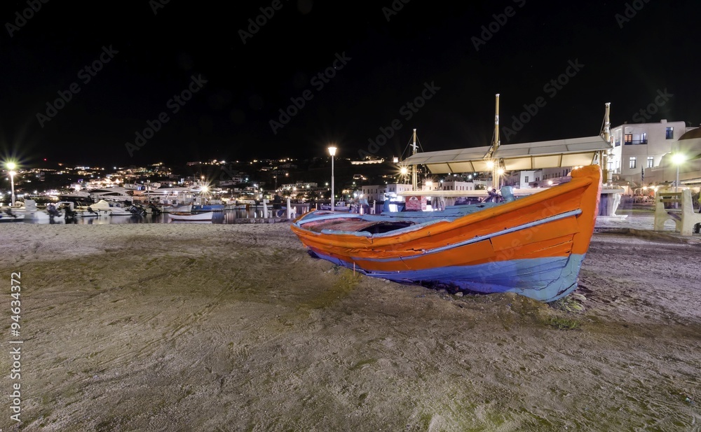 Night view of Chora port in Mykonos, Greece. Hora town cityscape lights reflected on the sea, a typical orange, blue greek island fishing boat in the harbour sandy shore and whitewashed buildings.