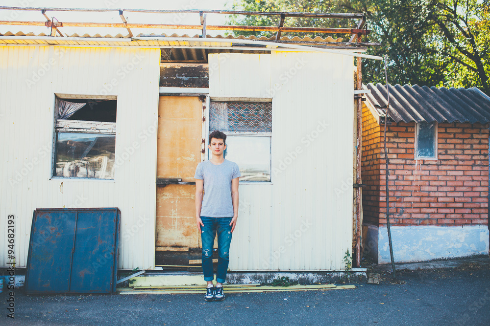 Young man standing still on destroyed house background. Teenage hipster wearing casual wear posing for camera near abandoned building Young man standing still on destroyed house background. Teenage hipster wearing casual wear posing for camera near abandoned building