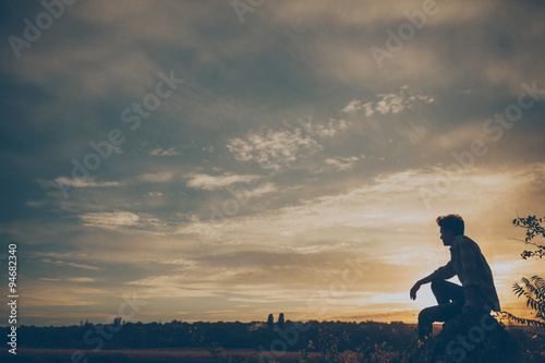 Silhouette of young man sitting on sunset or sunrise. Confident teenage boy thinkig on cliff stone. Hope. Sadness. Freedom. © kegfire