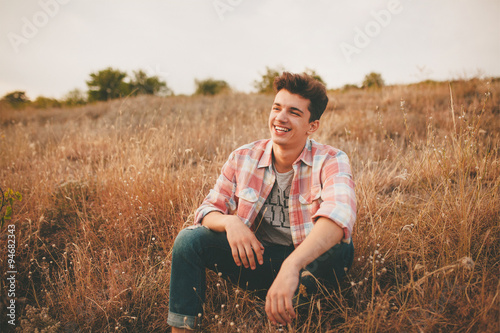 Handsome teenage boy wearing casual shirt sitting on grass on autumn day. Happy smiling young man sitting outdoors.