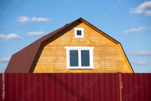 New suburban rural wooden house painted brown with white plastic windows after red metal fence on sunny day © DyMaxFoto