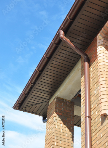 New Rain Gutter Drain Pipe Downspout Installation on the Unfinished House Facade Brick Wall Outdoor. Install Drainage System with Plastic Siding Soffits and Eaves Exterior. photo