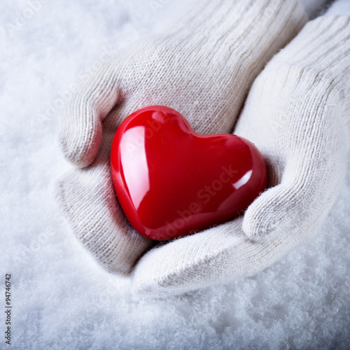 Female hands in white knitted mittens with a glossy red heart on a snow background.  Love and St. Valentine concept