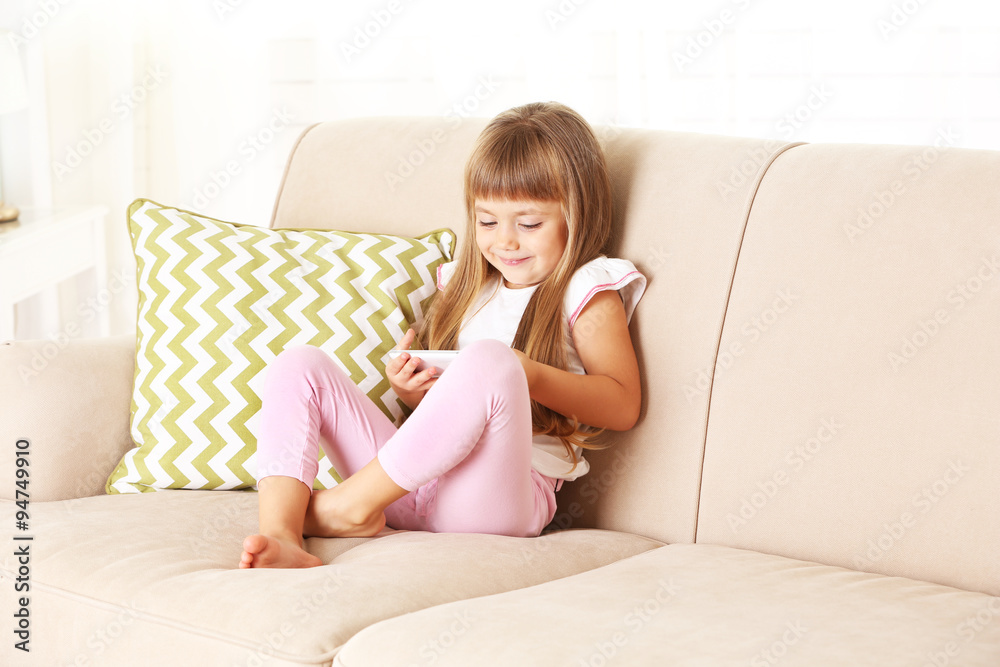 Beautiful little girl with smart phone sitting on sofa, on home interior background Beautiful little girl with smart phone sitting on sofa, on home interior background