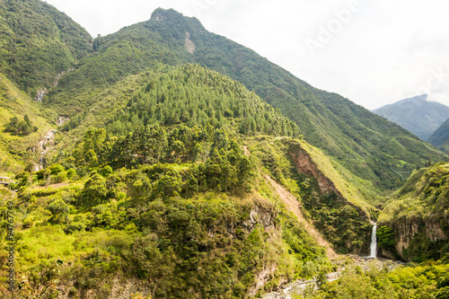 A breathtaking view of a verdant mountain valley with dense green foliage on steep slopes and a slender waterfall tumbling down a rocky cliffside near Banos in the Andes of Ecuador.