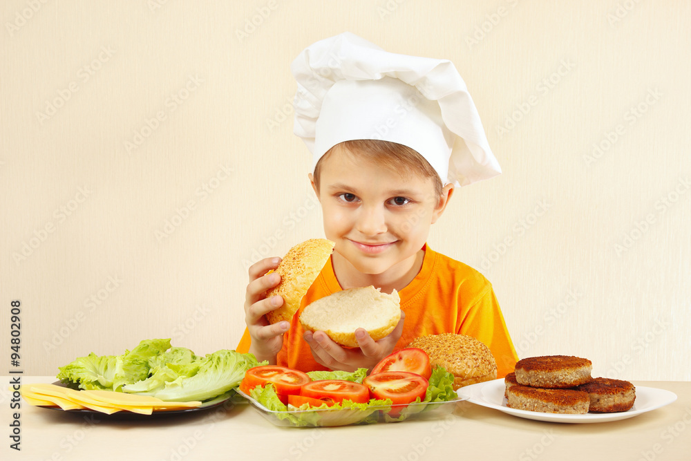 Little funny chef at the table with ingredients is going to cook a hamburger Little funny chef at the table with ingredients is going to cook a hamburger