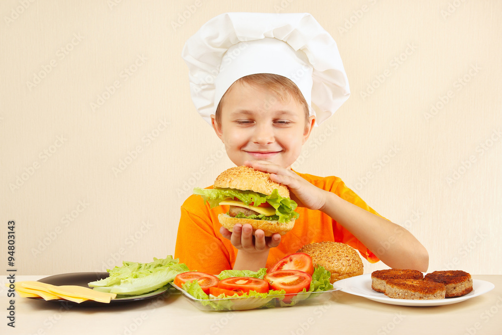 Little funny chef in chefs hat enjoys a cooking tasty hamburger Little funny chef in chefs hat enjoys a cooking tasty hamburger