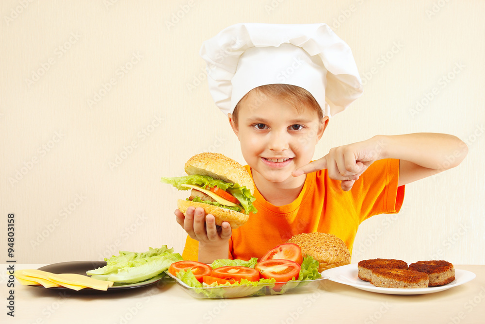 Little boy in chefs hat expressive enjoys a cooked hamburger Little boy in chefs hat expressive enjoys a cooked hamburger