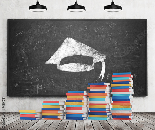 A stair is made of colourful books. A graduation hat is drawn on the black chalkboard. Concrete wall, wooden floor and three black ceiling lights. photo