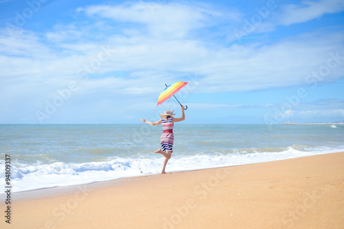 Beautiful young excited woman with rainbow umbrella dancing on sunny seashore