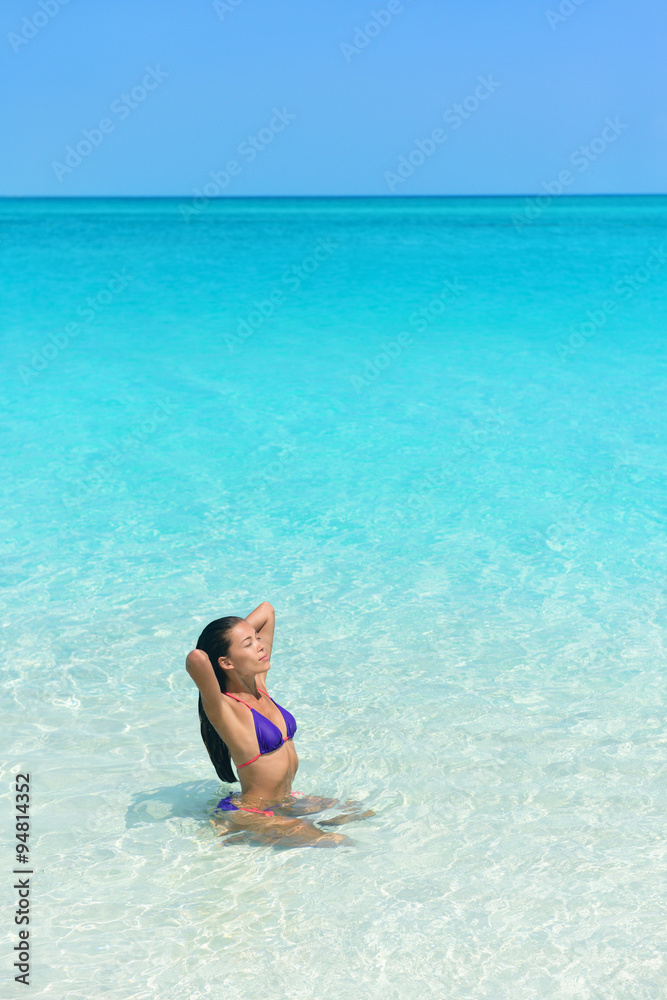 Beach woman in bikini swimming in blue ocean. Beautiful Asian girl sun tanning and relaxing in water taking care of her hair and skin.