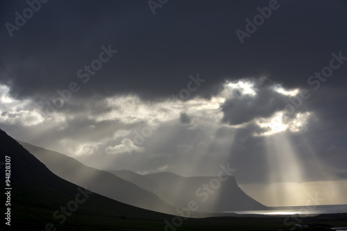 Fototapeta Naklejka Na Ścianę i Meble -  Küste in den Westfjorden von Island,dunkle Wolken an Islands Küste. Sonnenstrahlen durchbrechen die Wolkendecke.Ruhe vor dem Sturm