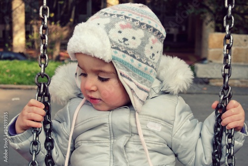 Portrait of a little girl on the swing in a park, wearing warm winter clothes and a white cap. Image filtered in faded, retro, Instagram style; nostalgic, vintage childhood concept.