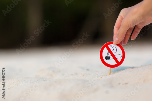 Female hand holding "No smoking" sign on the beach on white sand background