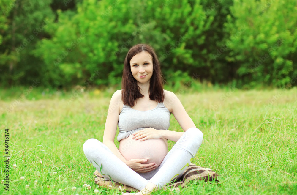 Happy smiling pregnant woman sitting on grass doing yoga in summ