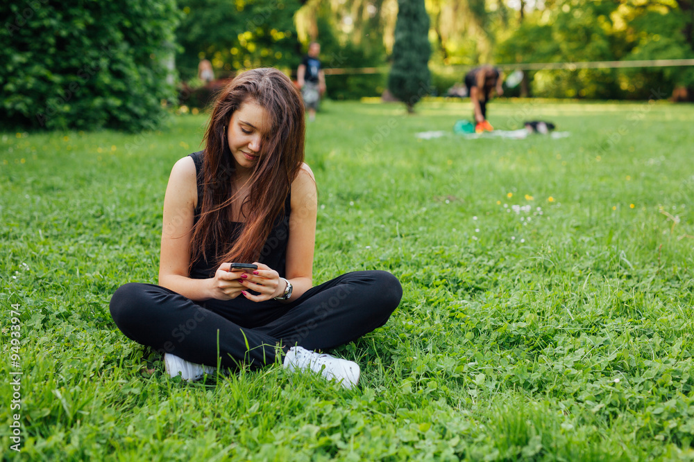 Brown hair girl sitting in the park on the grass and using smartphone.