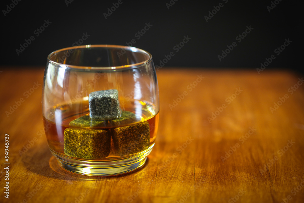 A glass of whiskey on the rocks in simple glass ware with whiskey stones on top a wood desk.