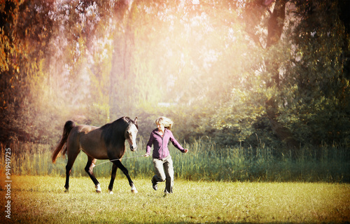 Woman and brown horse running across meadow with big trees