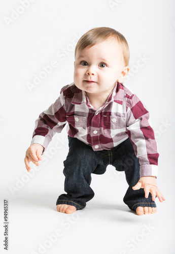 a cute 1 year old baby stands in white studio with jeans and a red white flannel looking camera left in excitement