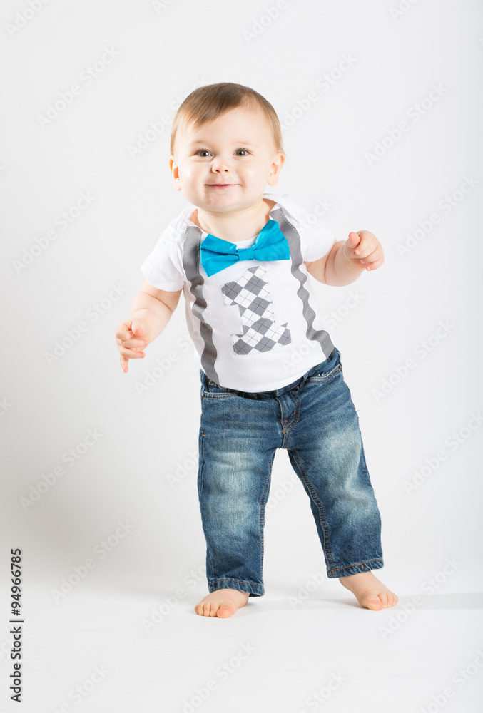 a cute 1 year old stands in a white studio setting. The boy has a happy expression. He is dressed in Tshirt, jeans, suspenders and blue bow tie a cute 1 year old stands in a white studio setting. The boy has a happy expression. He is dressed in Tshirt, jeans, suspenders and blue bow tie
