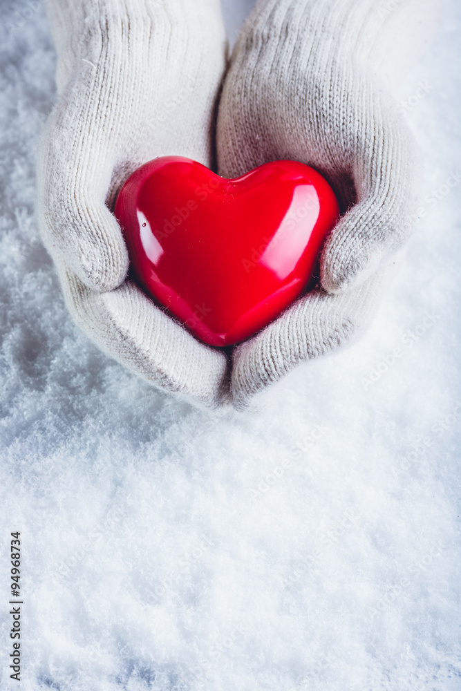 Female hands in white knitted mittens with a glossy red heart on a snow background. Love and St. Valentine concept.