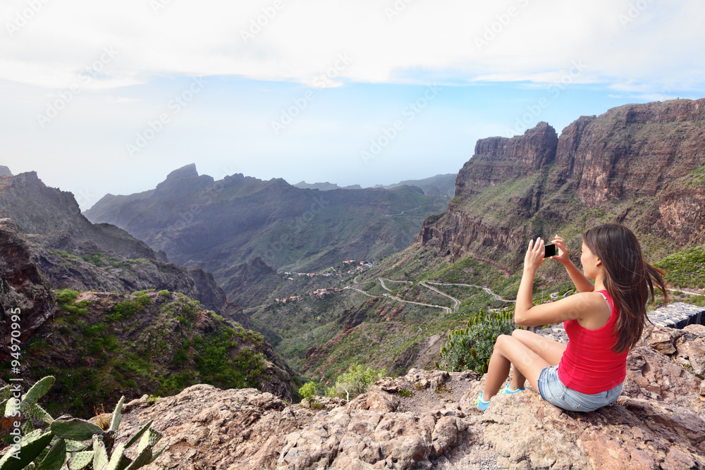 Woman photographing mountains while sitting on cliff. Young female is using smart phone to click picture of beautiful nature. Rear view tourist visiting Tenerife, Masca Valley, Canary Islands, Spain.