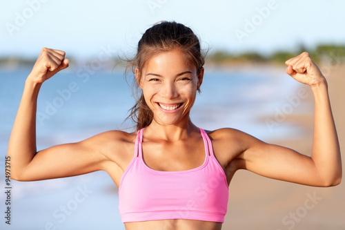 Happy sporty fitness woman flexing muscles on beach. Smiling young is wearing pink sports bra. Female is showing her strength and healthy lifestyle on sunny day.