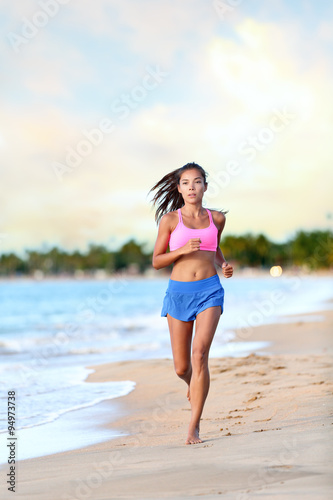 Confident young woman jogging on beach against sky. Full length of determined female is in sports clothing. Jogger is exercising at sea shore during sunny day.