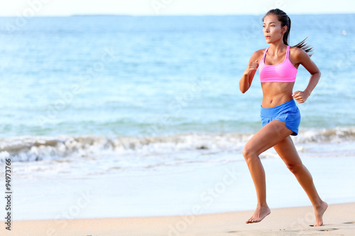 Running Determined young woman runner jogging on beach. Full length of fit female is in sports clothing. Jogger is exercising at sea shore during sunny day.