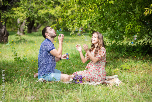 Happy young couple expecting baby, pregnant woman with husband blowing bubbles and laughing, sitting on green grass © Andrey Bandurenko