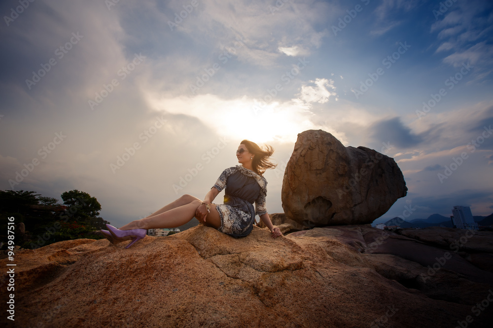 brunette girl in short grey frock sits on rock against sky