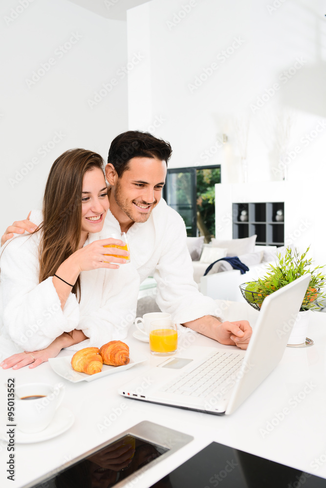 young couple in bathrobe at home having coffee in the kitchen and working on laptop computer
