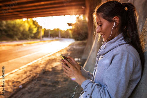 Young woman relaxing after jogging.She using her smart phone and listening to music.Typing message.leaning against wall. photo